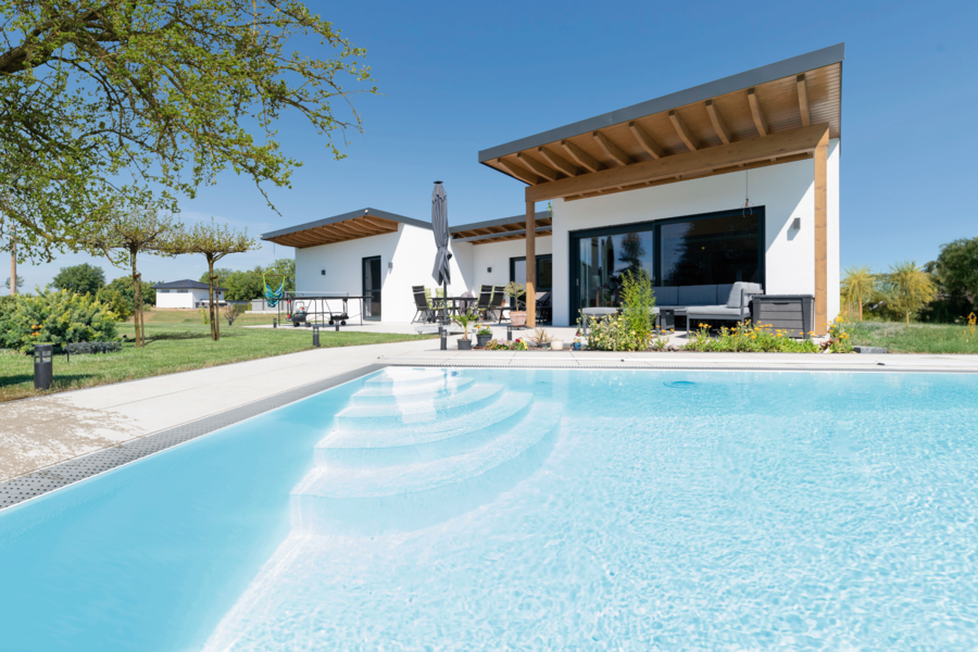 Hartl Haus zeigt ein weisses Einfamilienhaus mit Flachdach und einer großen Terrasse mit überdachten Bereich und gemütlichen Loungemöbeln und einem rechteckigen Pool im gepflegten Garten.
