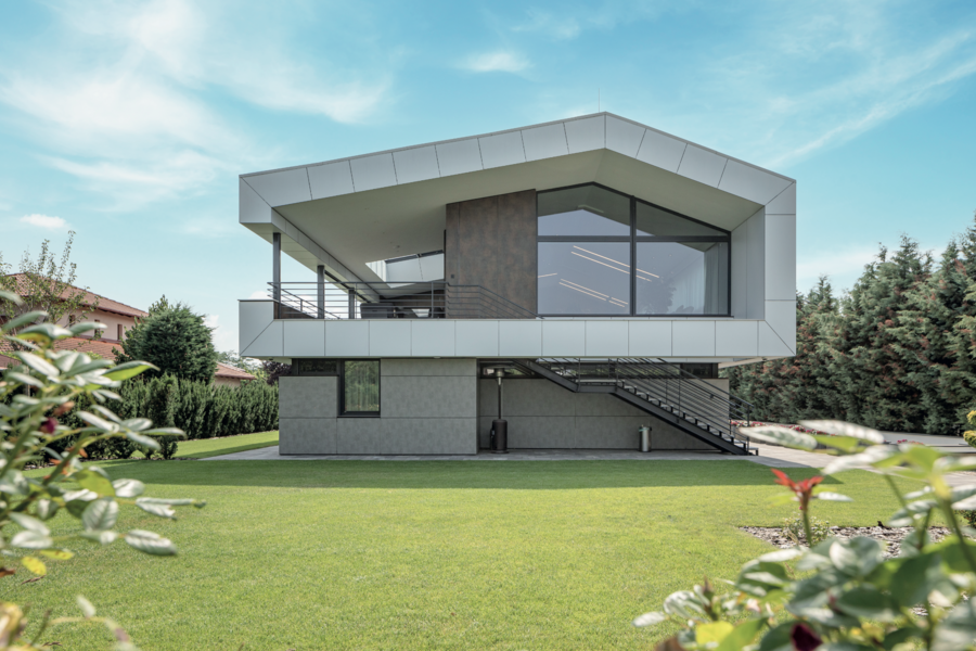 Internorm zeigt ein graues Haus mit weisser Verkleidung beim Dach, großen Fenstern und Steinplatten als Weg um das Haus, mit hellen Treppen und Geländer.