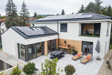 Internorm zeigt ein weisses Haus mit dunkler Photovoltaikanlage am Dach, einer Terrasse mit Steinplatten, Holzliegen und einem schwarzen Esstisch mit Stühlen.