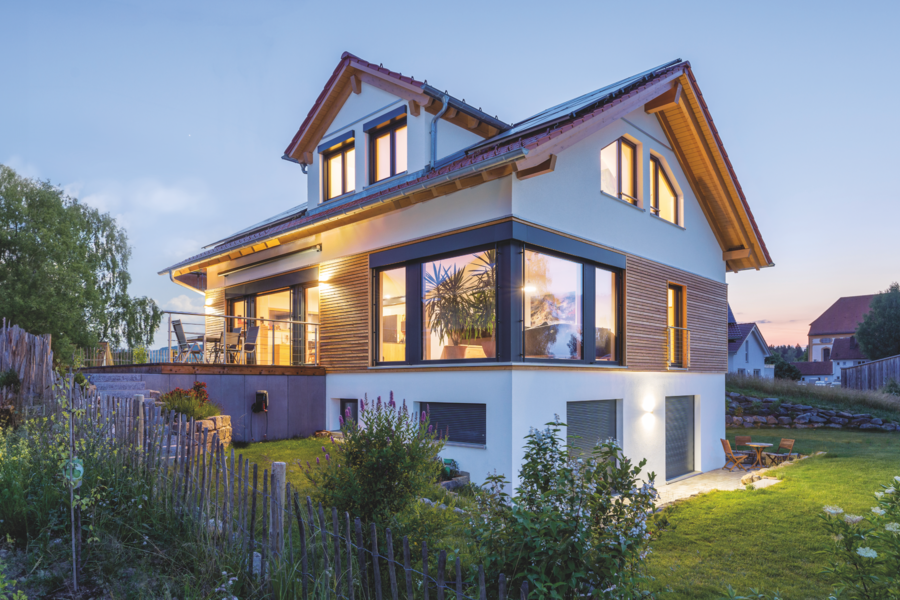Regnauer zeigt ein weisses Einfamilienhaus mit Holzakzenten, großen Fenstern mit schwarzen Rahmen, einer Terrasse mit einer Treppe die zu einem gepflegten Garten führt.