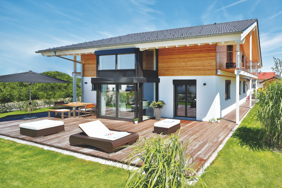 Regnauer zeigt ein Einfamilienhaus in weiss und mit einer hellen Holzverkleidung, einem Balkon und einer großen Terrasse mit Holzdielen und gemütlichen Outdoormöbeln.