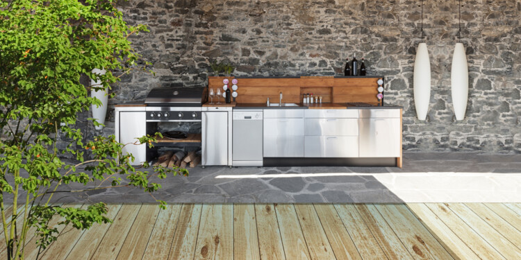 Moderne Outdoorküche mit Elementen aus Holz und Nirosta-Edelstahl überdacht und witterungsgeschützt an einer Natursteinwand.