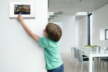 ABB - Busch-Jäger zeigt einen kleinen Jungen, welcher an Wandsteuerung mit Kamera tippt im Esszimmer.