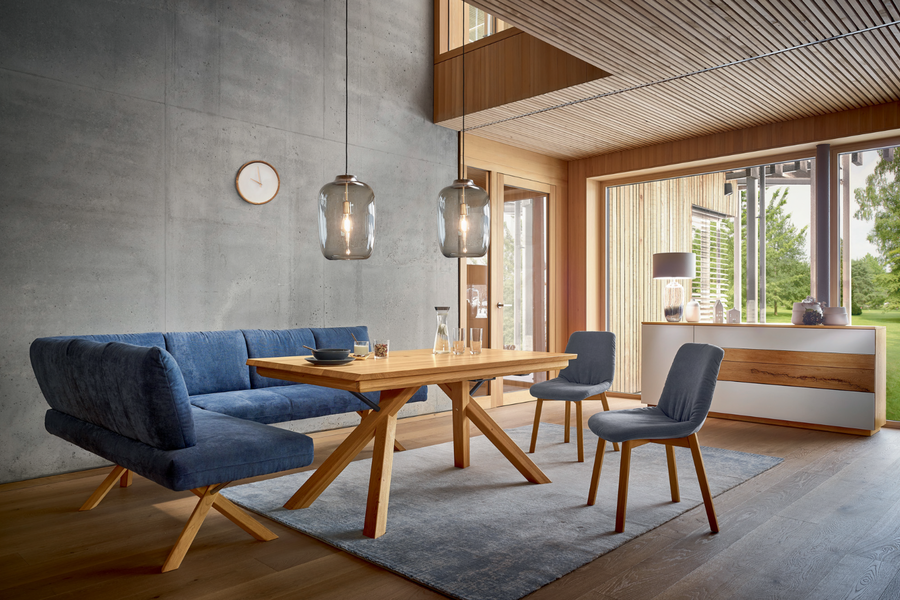 BÖHM MÖBEL zeigt einen Essbereich mit zwei blauen Stoffstühlen, einer Sitzbank, einem Holztisch, zwei zylinderförmige Hängelampen und einem grauen Teppich auf dem Holzboden.
