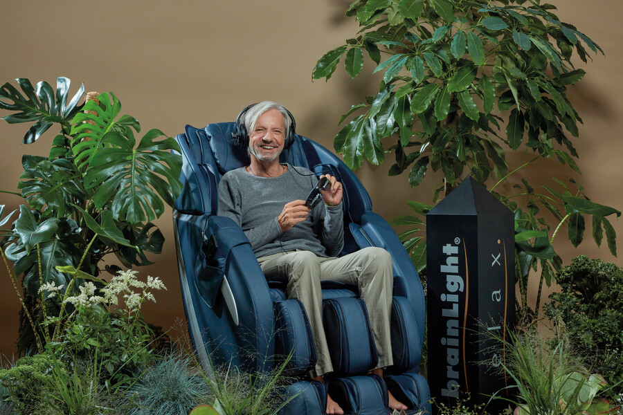Ein älterer Herr mit Kopfhörern, lässt sich von dem dunkelblauen Massagestuhl von brainLight, in einer bepflanzten Entspannungsoase massieren.