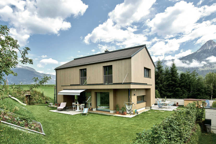 ERLER BAU GmbH zeigt ein modernes Einfamilienhaus aus Holz mit großen Fenstern und einer mit Markise überdachten Terrasse mit Fliesenboden und gemütlichen Möbeln im gepflegten großen Garten.