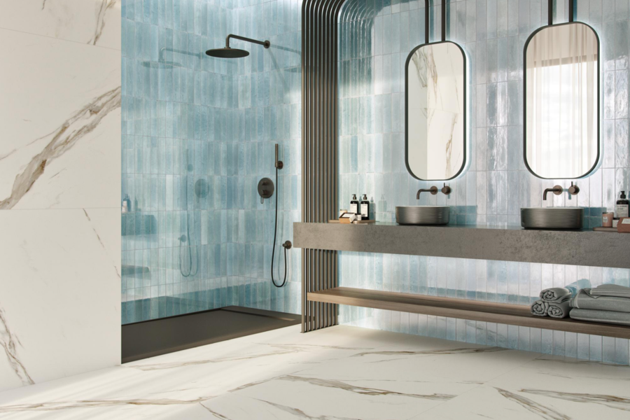 Fliesendorf zeigt ein Badezimmer mit offener Dusche, Doppelwaschtisch mit großen Spiegeln und dunklen Waschbecken, blauen Wandfliesen und einen marmoriertem Fliesenboden.