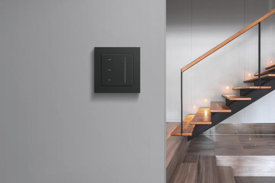 Gira zeigt eine schwarze Lichtsteuerung an einer grauen Wand im Treppenhaus mit Holztreppen und einem Geländer aus Glas mit einer Haltestange aus Holz.
