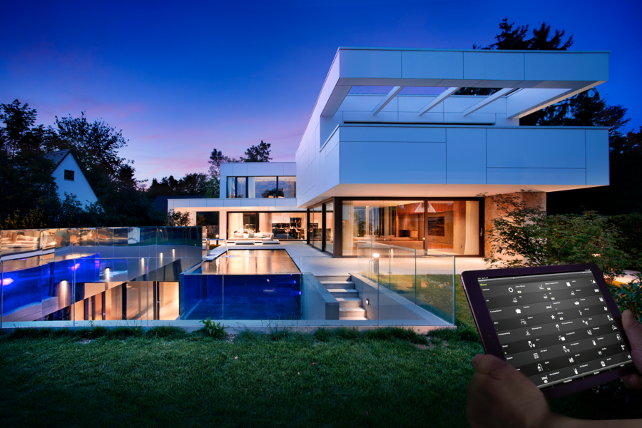 Gira zeigt eine moderne Villa mit Flachdach, Pool im Garten und einem Smarthome Tablet für die automatische Steuerung der Beleuchtung.