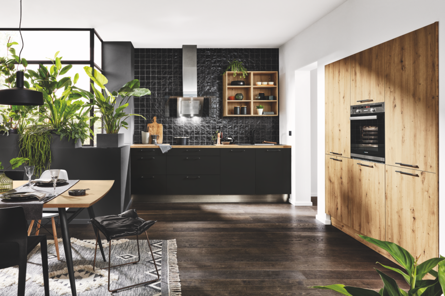 WOHNHAUS Grill & Ronacher zeigt eine schwarze Häcker Küche mit einem Wandschrank aus hellem Holz und anschließender Essgruppe mit einem Holztisch und schwarzen Stühlen.