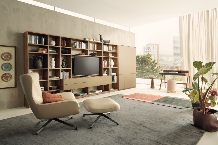 WOHNHAUS Grill & Ronacher zeigt einen Wohnbereich mit einem Fernsehsessel mit Hocker aus Leder und einem großen Wandverbau mit großem Fernseher und einem Schreibtisch vor dem Fenster von Anrei.