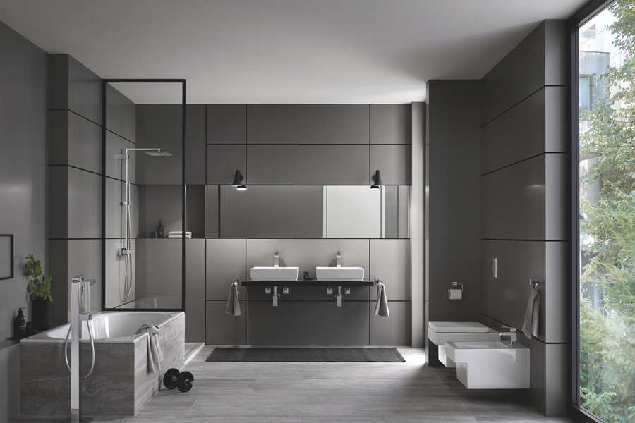 GROHE zeigt ein Badezimmer mit grauen Wandfliesen, freiliegendem Teppich, Toilette, Bidet, Doppelwaschtisch und Badewanne mit freistehender Armatur sowie einer Duschkabine mit Glaswand.