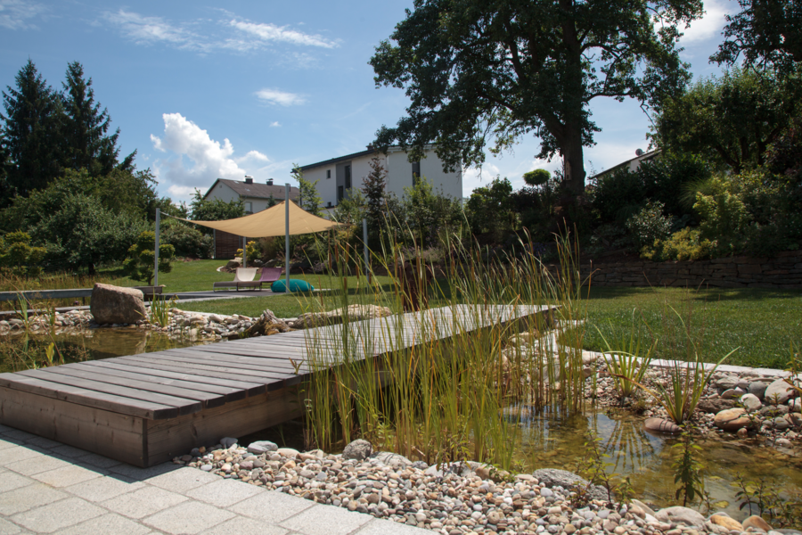 Ein Sonnensegel sorgt für effizienten Sonnenschutz am Pool und dem kleinen Teich mit Holzsteg von Hablesreiter.