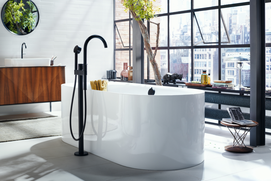 Hansgrohe präsentiert eine ovale, weisse Badewanne mit schwarzer Armatur in einem durch die großen Fensterflächen sehr hellen Badezimmer.