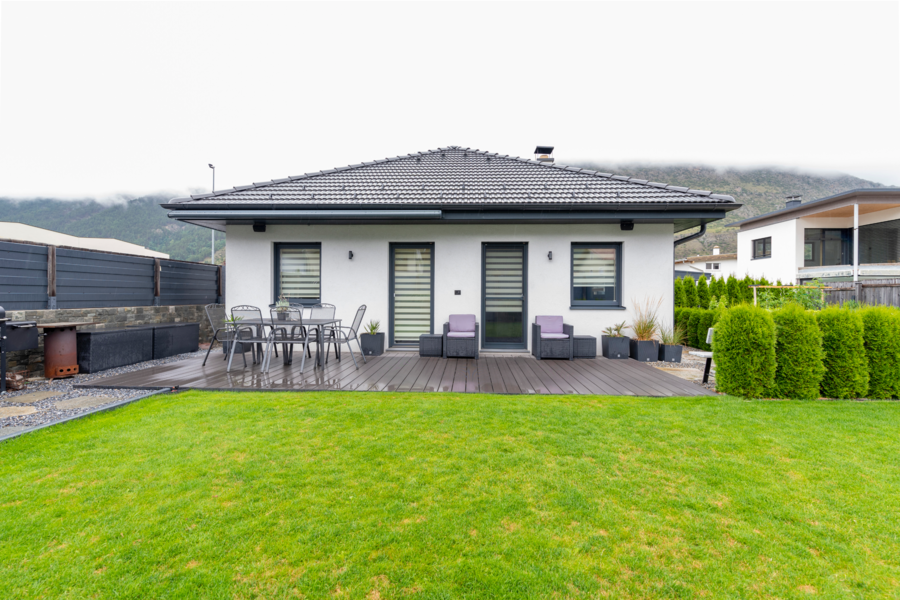 Hartl Haus zeigt ein weißes modernes Haus mit schwarzem Walmdach, einer Terrasse mit Holzdielen am Boden, Gartenmöbeln und einem dunkelgrauen Sicht- und Lärmschutzzaun.