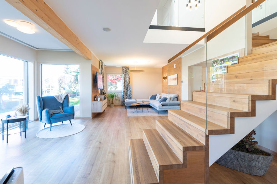 Hartl Haus zeigt ein Wohnzimmer mit Holzboden, einem blauen Couchstuhl, Treppenaufgang in den ersten Stock und großen Fensterflächen.