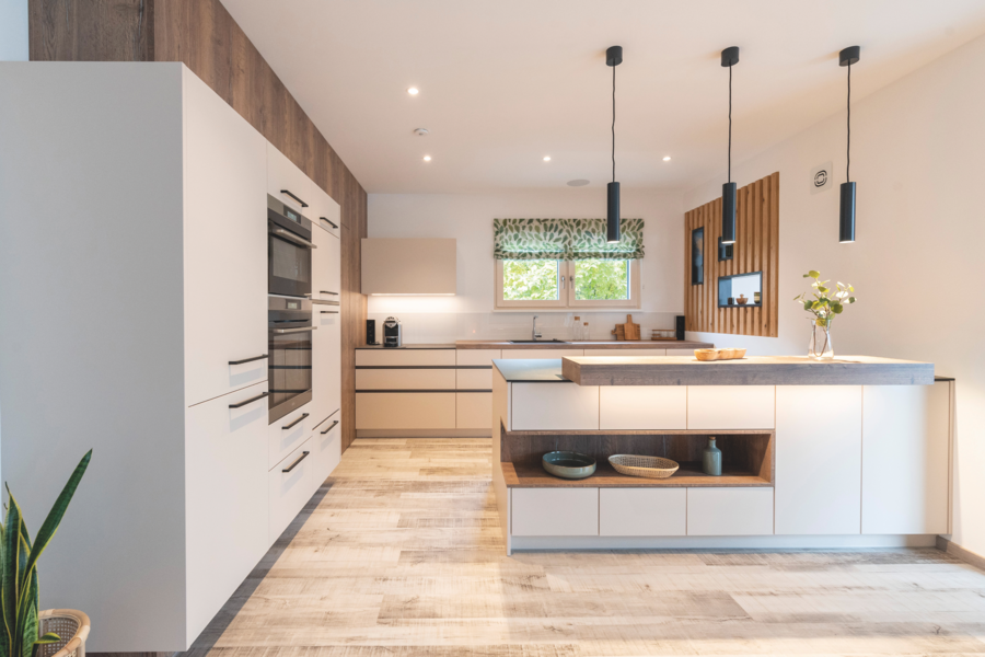 Hartl Haus zeigt eine Küche mit weissen Fronten, Holzelementen und viel Stauraum mit schwarzen Hängelampen und einem hellen Boden.