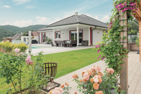 Hartl Haus zeigt einen weissen Bungalow mit überdachter Terrasse, gefliestem Boden und einer Sitzgruppe aus Holz, schönen Blumen und Kletterpflanzen.