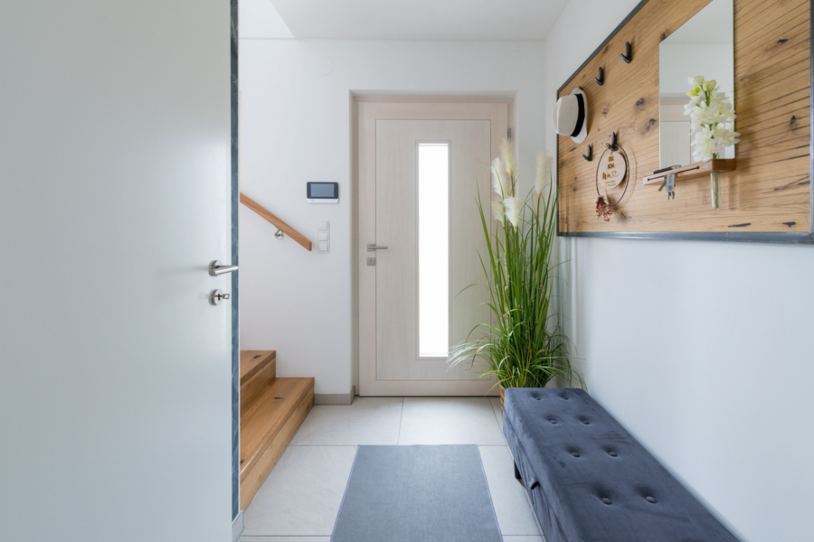 HARTL HAUS zeigt den minimalistischen Eingangsbereich mit niedrigen Bänkchen, weißer Haustür mit Glasstreifen und Holztreppe.