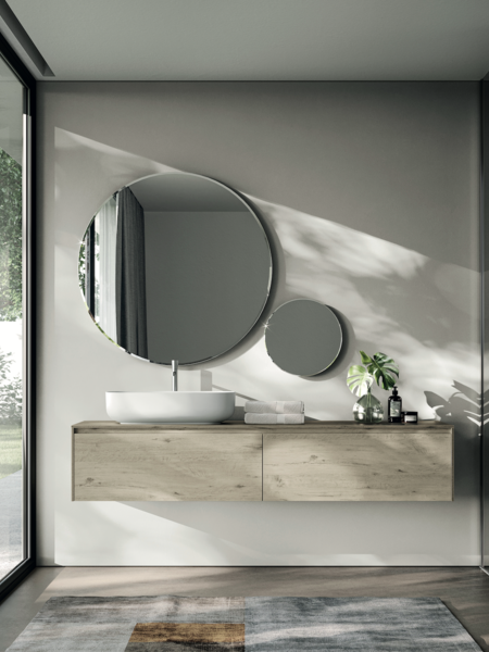 IDEA GROUP zeigt ein Badezimmer mit runden Spiegeln, einen Waschtisch aus Holz und einem weissen Waschbecken mit silbernen Armaturen und Fenster mit Blick in den Garten.