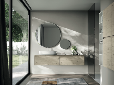 IDEA GROUP zeigt ein Badezimmer mit runden Spiegeln, einen Waschtisch aus Holz und einem weissen Waschbecken mit silbernen Armaturen und Fenster mit Blick in den Garten.