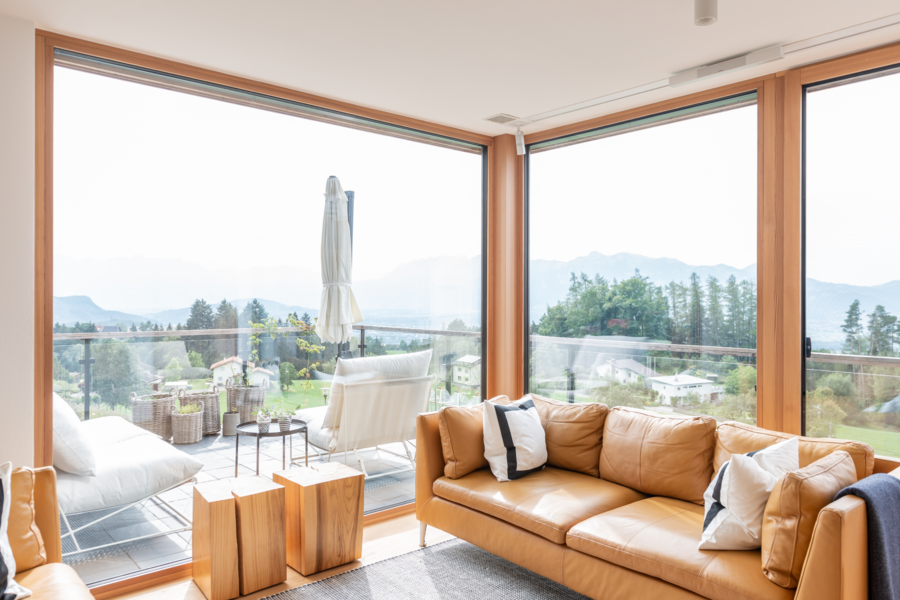 Internorm zeigt ein lichtdurchflutetes Wohnzimmer mit Glasfronten und einer braunen Ledercouch mit Blick ins Grüne und Zugang zum Balkon mit Loungemöbeln.