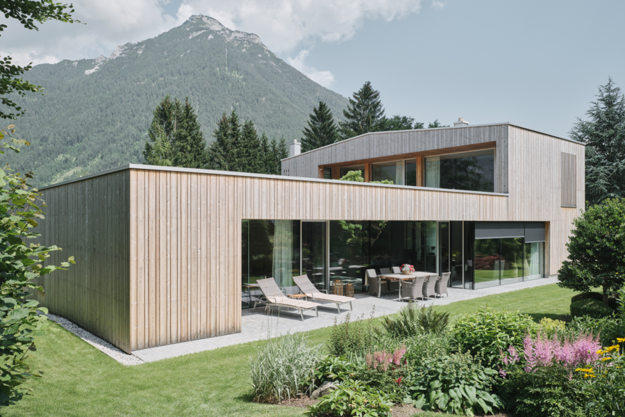 Josko zeigt ein modernes Einfamilienhaus mit Flachdach, einer hellen Holzverkleidung und sehr großen Glasfronten und einer Schiebetür aus Glas zur überdachten Terrasse.