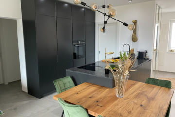 Einrichtung & Design Köck zeigt eine Referenzküche von Intuo in schwarz mit inkludiertem Esstisch aus Holz und grünen Stühlen aus Samt.