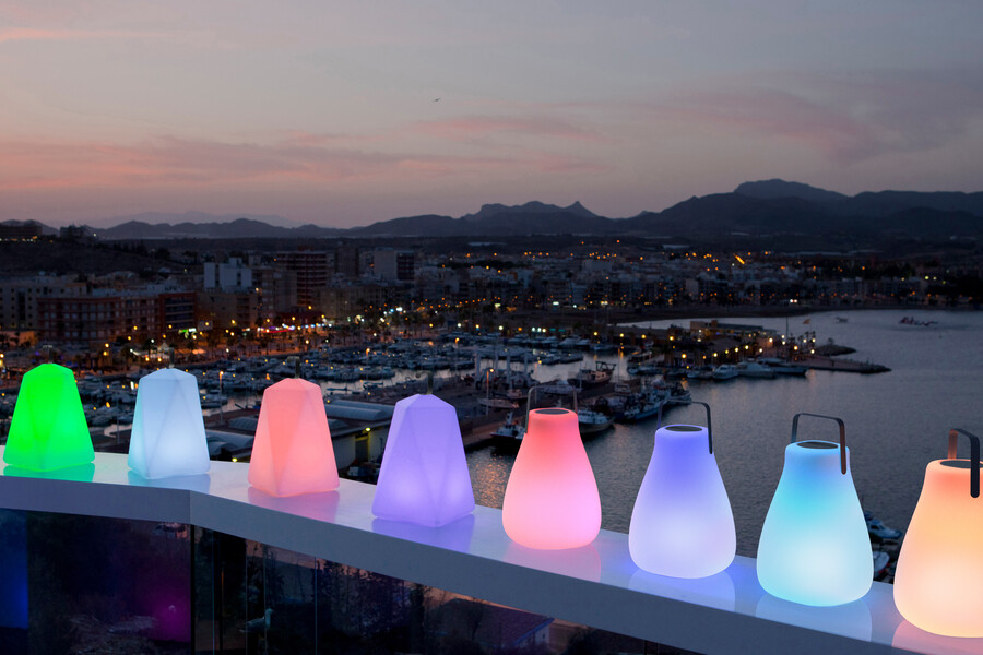 Lampenwelt präsentiert die neuen Solarlampen in buntem Design auf einem Glasgeländer vor einer Stadt mit Hafen.