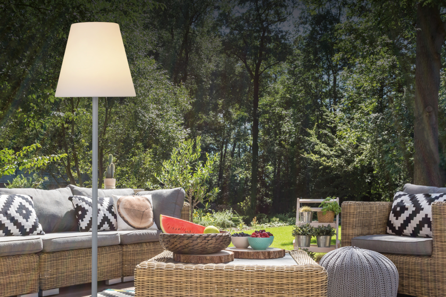 Lampenwelt präsentiert die neuen Solarlampen  als Stehlampe neben einer gemütlich aussehenden Rattanlounge mit Obst am Tisch und Wald im Hintergrund.