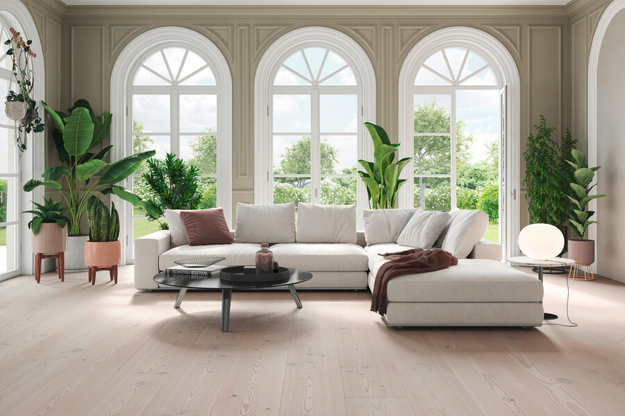 Ein Wohnzimmer in einem sehr hohen Raum mit hellgrauer L-Couch, großen Fensterflächen und mehreren Pflanzen, geschmückt von einem mafi Naturholzboden.