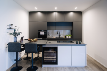 MF Interieur zeigt eine Küche mit großem, grauen Einbauschrank und einer weissen Kücheninsel mit Kochfeld, einem Weinkühler und dunkler Bar mit Hockern.