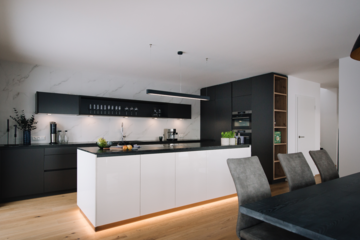 MF Interieur zeigt ein helles Esszimmer mit dunklem Holztisch und hellgrauen Stühlen und eine Küche mit weissen Fronten und schwarzen Schränken mit einer Kücheninsel.