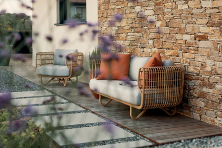 Hausfassade aus braunem Stein mit schmaler Holzterrasse neben der Hausmauer, ausgestattet mit modischen Gartenmöbeln - Garten gestaltet von Nentwich Gartenbau.