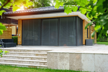 Newo Sonnen- und Insektenschutz zeigt ein Poolhaus mit Flachdach und Holzverkleidung an dessen Terrassentüren schwarze Rollläden montiert sind und eine geflieste Terrasse deren Stufen in den Garten führen.