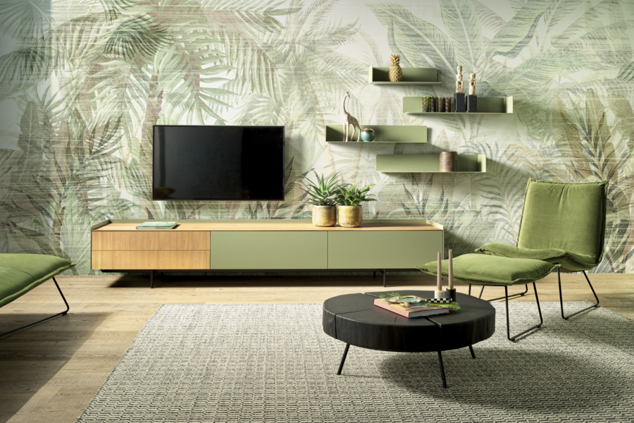 Pointinger wohnen zeigt ein Wohnzimmer mit grünen Palmen-Tapeten, grünen Hängeregalen, einem Sideboard mit TV, hellem Teppich, Couchtisch und grünem Sessel von haas.
