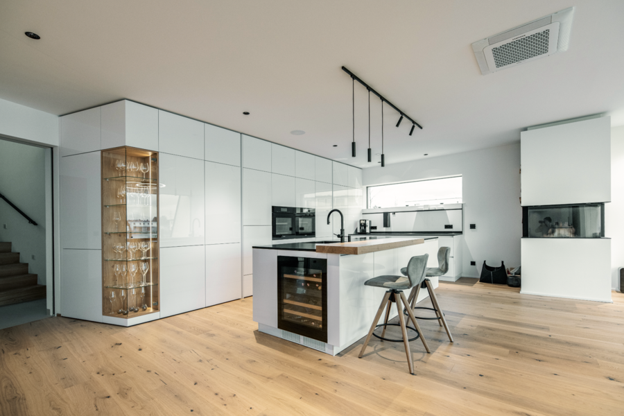 reginaplaza zeigt eine moderne Küche in weiss Hochglanz  und einer Kochinsel mit Kochfeld, Spülbecken und mit einer Arbeitsplatte aus Holz und einem Weinkühlschrank in schwarz.