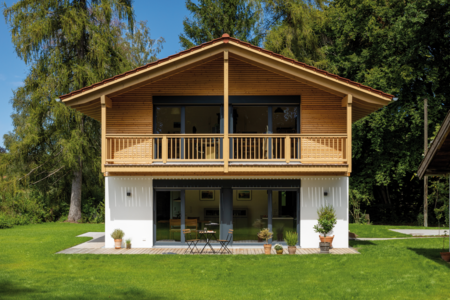 Regnauer zeigt ein Einfamilienhaus im Grünen mit schöner Holzverkleidung, einem Balkon mit Holzgeländer und einer überdachten Terrasse mit Holzboden und Bistro Möbeln. 