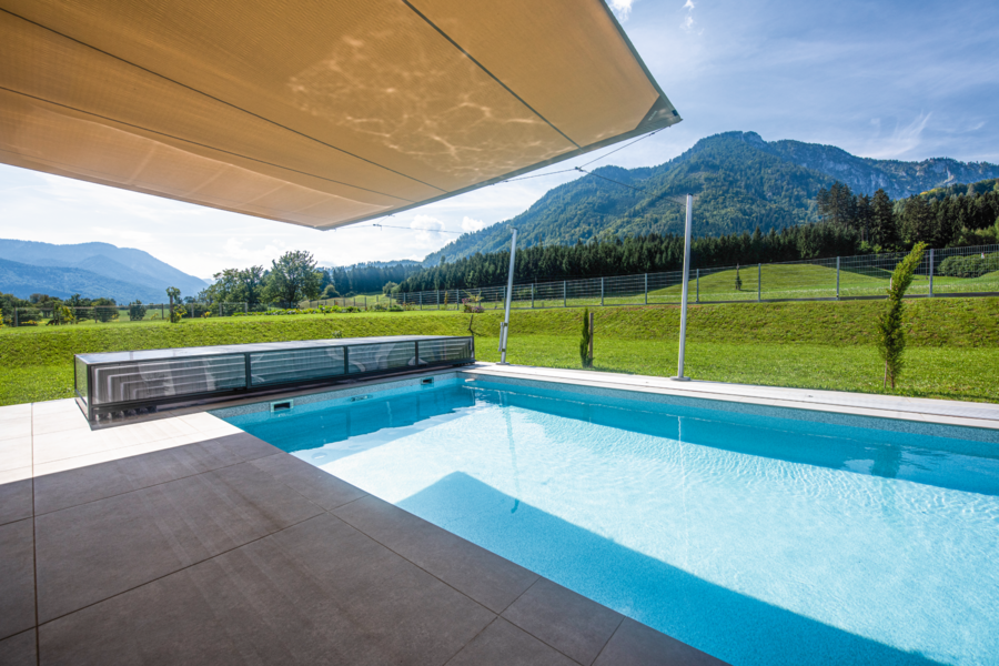 RUHA zeigt einen rechteckigen Pool mit einer Poolüberdachung aus Glas und einer gefliesten Poolumrandung in einem großen Garten mit gepflegtem Rasen.