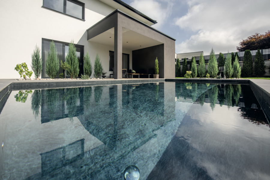 RUHA zeigt einen edlen Pool der im Boden versenkt ist und eine geflieste, graue Umrandung hat im gepflegten Garten von einem Einfamilienhaus mit überdachter Terrasse.