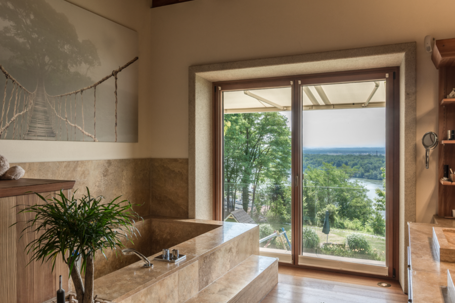 Silber Fenster zeigt ein elegantes Badezimmer mit braunem Naturstein und einer großen Terrassentür mit Blick ins Grüne.