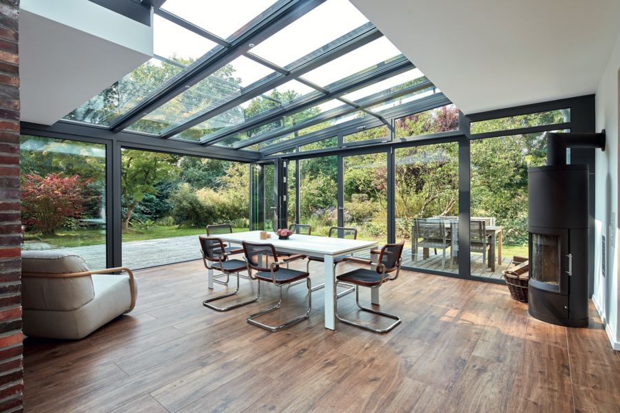 Solarlux zeigt einen Wintergarten mit großen Glasfronten, Panoramadach und einer Glasfaltwand mit einem weissen Esstisch und modernen Stühlen aus Materialmix.