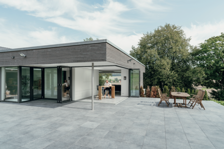 Solarlux zeigt eine geflieste Terrasse mit überdachtem Bereich mit einem Hochtisch und rustikaler Sitzgruppe aus Holz und eine Glasfaltwand mit Zutritt zum Wohnzimmer.