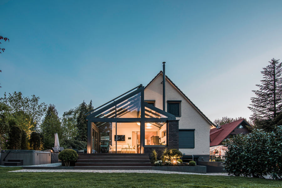 Solarlux zeigt ein modernes Einfamilienhaus mit großem, verglasten Wintergarten mit gemütlicher Sitzlandschaft und Glasschiebetüren mit Zugang zum Garten mit Pool und gefliester Terrasse.