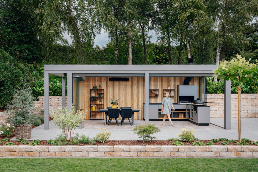 Solarlux zeigt einen Garten mit kleiner Ziegelmauer und überdachter Terrasse mit Holzverkleidung an der Rückwand und Verglasung, mit einer Outdoorküche und gemütlicher Sitzgruppe. 