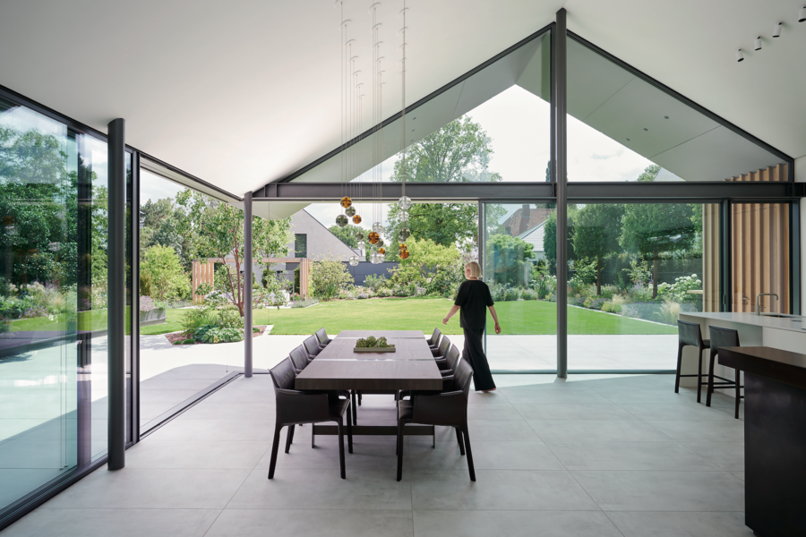 Solarlux zeigt ein offenes Esszimmer mit Küchenzeile und großem Esstisch mit Lederstühlen und Zugang zum gepflegten Garten mit gefliester Terrasse durch mehrere Glasschiebetüren.