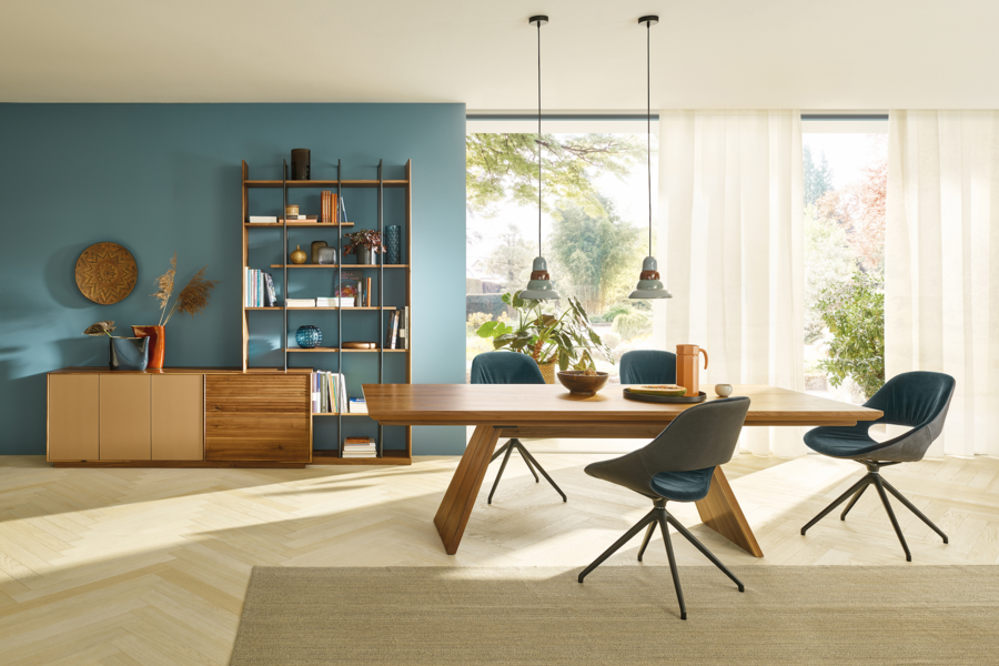 Tischlerei Bauer zeigt ein Esszimmerr mit einem Holztisch und blauen Stühlen auf einem hellen Teppich, eine Wandkommode und Blick in den Garten von Anrei.