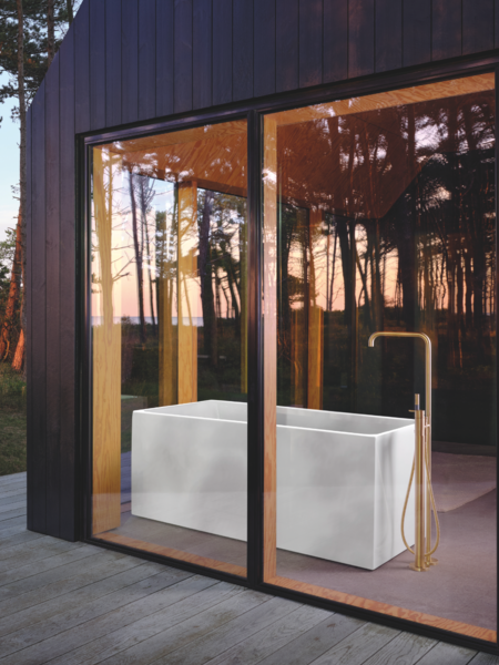 VOLA zeigt eine Waldhütte aus Holz mit großen Fenstern und einer freistehenden Badewanne mit goldenen Armaturen.