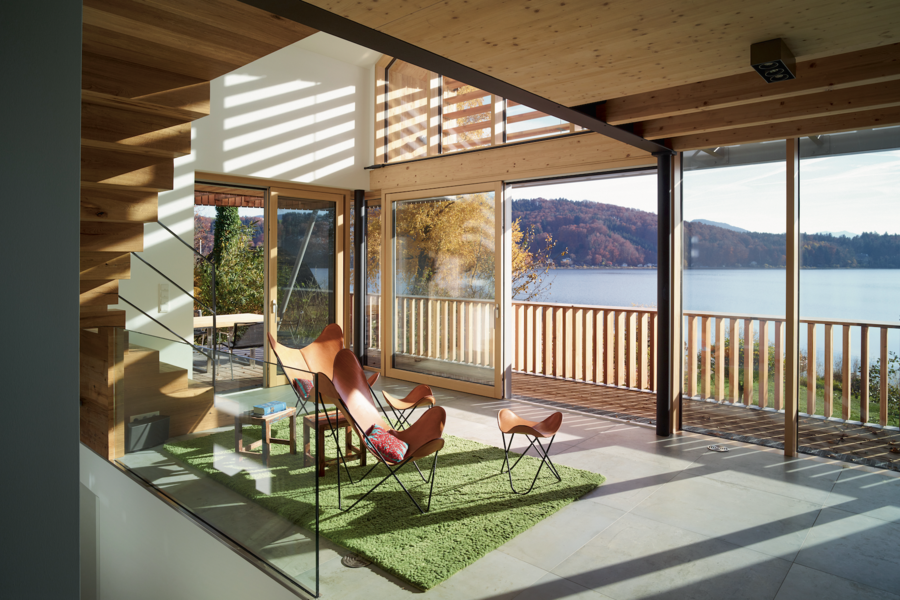 Weissenseer Holz-System-Bau zeigt einen lichtdurchfluteten Loungebereich mit Ledersesseln und grünem Teppich mit Zugang zum Balkon durch Glasschiebetüren in einem modernen Holzhaus.