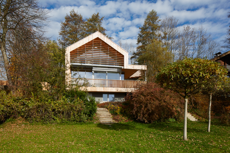 Weissenseer Holz-System-Bau zeigt ein schönes, modernes Holzhaus mit überdachtem Balkon mit Holzgeländer und viel Bäumen rundherum und Steintreppen die zu einer großen Rasenfläche führen.